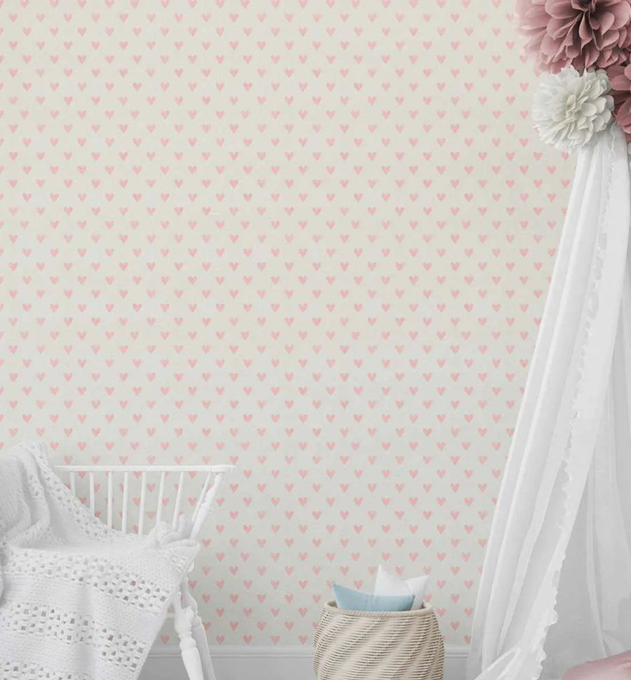Nursery:
Pink hearts wallpaper featuring a repeating pattern of small, hand-drawn hearts in a soft blush pink on a warm cream background, with a subtle textured finish. The wallpaper covers the entire wall of a nursery, which includes a white wooden crib with a white knitted blanket and a woven basket with blue and white pillows. A white canopy with pink and white paper flowers hangs on the right side of the wall.