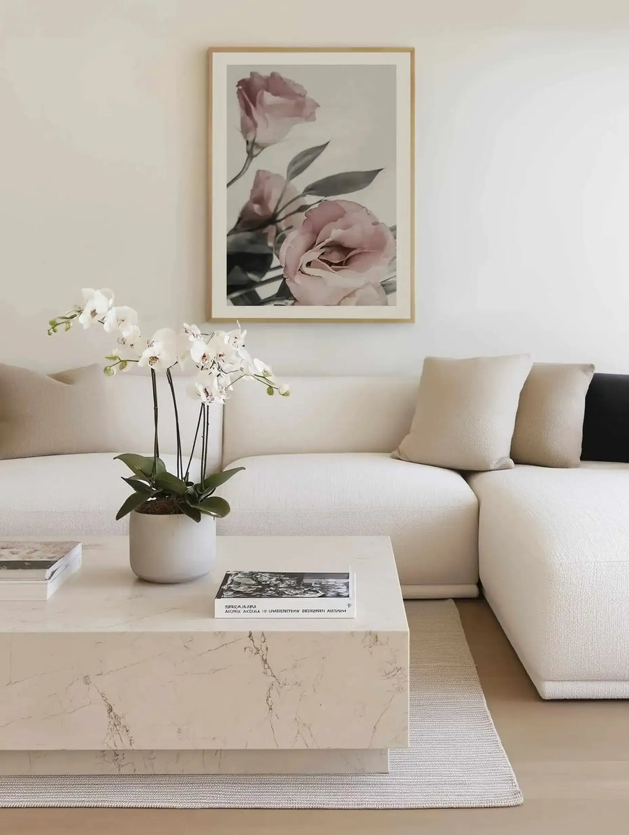 Framed art print featuring dusty rose and mauve lisianthus flowers with green foliage against a soft neutral background. The botanical photograph has a watercolor-like quality with muted tones. Natural wood frame with white border, displayed above a cream linen sectional sofa in a minimalist living room setting with marble coffee table and white orchid plant.