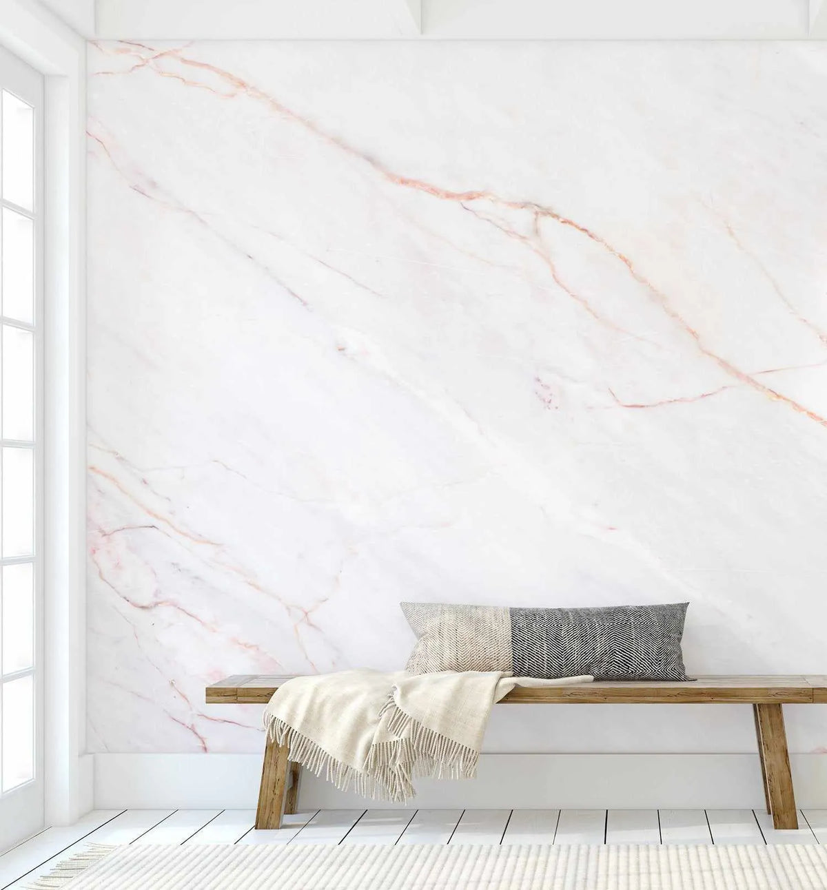 "Pink Marble peel and stick wallpaper displayed on a wall in a minimalist living room. The wallpaper features a repeating pattern of white marble with delicate, organic veins in soft rose pink and subtle grey, creating a luxurious and elegant backdrop. A light wood bench with a grey and cream patterned cushion and a cream throw blanket sits against the wall, with a white window frame visible on the left and a cream rug on the white plank floor."