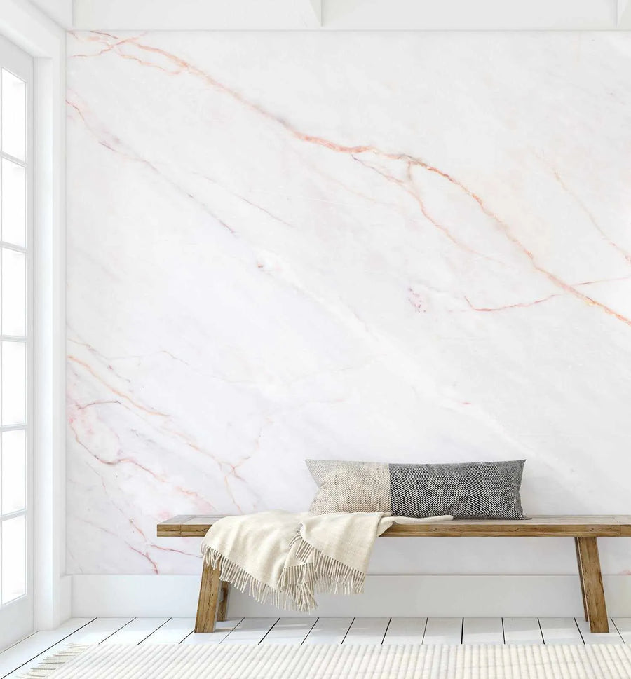 "Pink Marble peel and stick wallpaper displayed on a wall in a minimalist living room. The wallpaper features a repeating pattern of white marble with delicate, organic veins in soft rose pink and subtle grey, creating a luxurious and elegant backdrop. A light wood bench with a grey and cream patterned cushion and a cream throw blanket sits against the wall, with a white window frame visible on the left and a cream rug on the white plank floor."