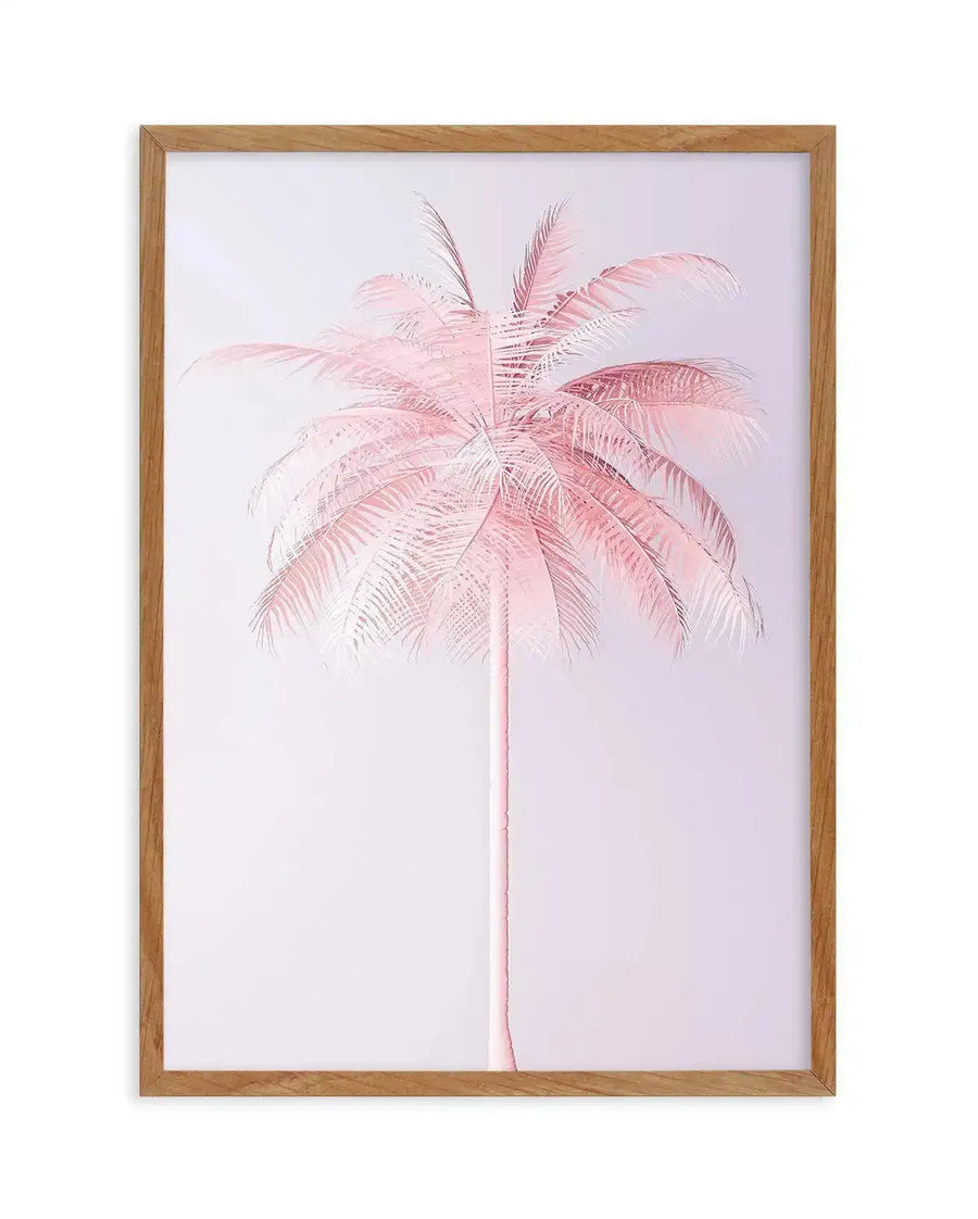 Framed art print featuring a single palm tree rendered in soft pink and mauve tones against a pale lavender background. The palm has a slender trunk and full, feathery fronds spreading outward in varying shades of blush pink and dusty rose with fine linear details. The artwork has a delicate, watercolor-like quality with subtle gradations of color. The print is displayed in a natural wood frame with a white border.