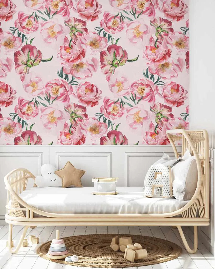 Lifestyle shot of a child's bedroom featuring peel and stick wallpaper with a repeating pattern of large-scale pink peony roses and buds with green leaves on a light pink background. The wallpaper covers the upper half of the wall above white wainscoting. A light wood rattan daybed with a white mattress is centered in the room, adorned with a white cloud pillow, a tan star pillow, and a white decorative drum. A round woven rug with wooden toys is on the floor.