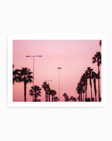 "Photography art print featuring a row of silhouetted palm trees against a vibrant pink and purple gradient sky at sunset. The composition includes streetlights with a warm glow, adding to the tropical and serene mood."