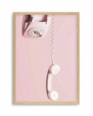 Framed art print featuring a vintage rotary telephone in a soft pastel pink, with the phone's body positioned upside down in the top left corner and the receiver hanging vertically downwards, connected by a coiled white cord. The background is a solid, light pink, and the print is displayed in a natural oak frame with a white border.
