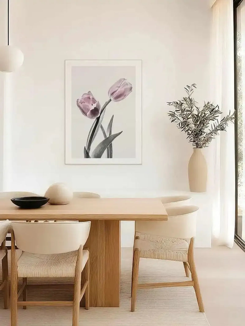 Framed art print displaying two dusty rose pink tulips with dark green stems and leaves against a soft beige background. The botanical illustration has a delicate, watercolor-like quality and is mounted in a light wood frame with a white border. The print is displayed on a white wall above a round wooden dining table in a minimalist interior with natural light from large windows, wooden chairs with cream cushions, and a cream ceramic vase containing dried greenery.