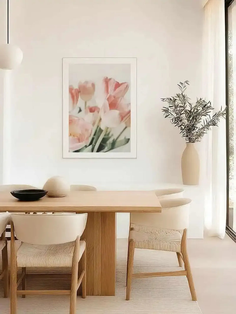 Lifestyle shot of a minimalist dining room featuring a framed art print of pink tulips. The print, displayed on a white wall above a light wood dining table, shows several soft pink tulips with green stems against a bright, slightly blurred background, suggesting a gentle, artistic photography style. The room includes light wood chairs with woven seats, a white pendant light, and a tall ceramic vase with olive branches next to a large window with sheer white curtains.