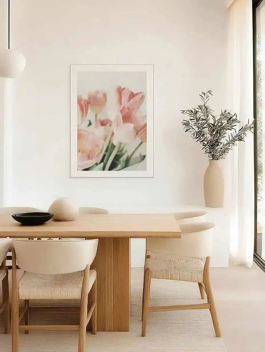 Lifestyle shot of a minimalist dining room featuring a framed art print of pink tulips. The print, displayed on a white wall above a light wood dining table, shows several soft pink tulips with green stems against a bright, slightly blurred background, suggesting a gentle, artistic photography style. The room includes light wood chairs with woven seats, a white pendant light, and a tall ceramic vase with olive branches next to a large window with sheer white curtains.