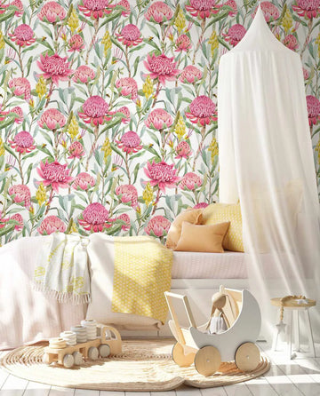 Lifestyle shot of a child's bedroom featuring Pink Waratah wallpaper. The wallpaper has a repeating pattern of hand-painted pink waratah flowers with green leaves and yellow flowering branches on a white background. The room includes a white bed with yellow and cream pillows and blankets, a white canopy, a light wood toy truck, and a white wooden toy pram with a stuffed bunny, all on a round woven rug.