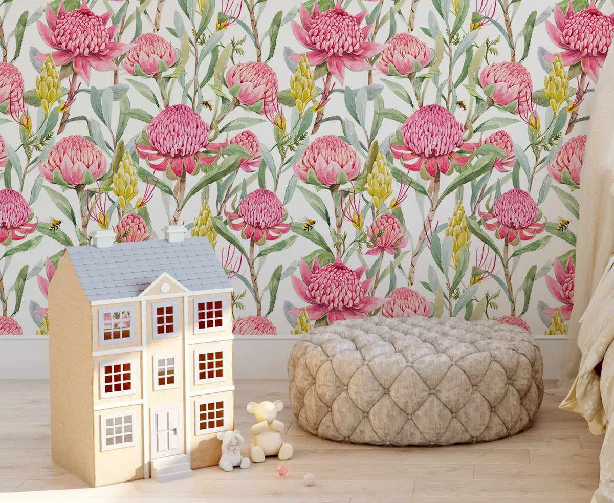 Lifestyle shot of a child's room featuring Pink Waratah wallpaper. The wallpaper has a repeating pattern of hand-painted Australian native flowers, including large pink Waratah blooms and yellow Banksia flowers, set against a white background with green foliage and small bees. A light brown wooden dollhouse with white trim and a grey roof sits on a light wood floor in the foreground, next to two small teddy bears and a quilted round ottoman.