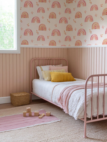 Lifestyle shot of a child's bedroom featuring a pink metal bed frame with white bedding and a pink throw, and a light pink wainscoting on the lower half of the walls. The upper half of the walls is covered in peel and stick wallpaper featuring a repeating pattern of hand-painted rainbows in shades of pink, orange, and cream, interspersed with small cross shapes and leaf motifs. The room includes a woven basket, a striped pink and cream rug, and wooden building blocks on the floor.