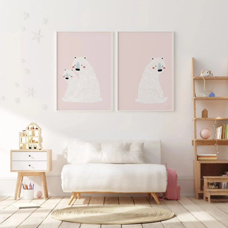 Lifestyle shot of a child's bedroom featuring two framed art prints above a white sofa. The left print depicts a mother polar bear and two cubs, while the right print shows a single polar bear, all illustrated with a soft, textured white fur, black noses, blue snouts, and pink cheeks on a pale pink background with a white frame and white border. The room has white walls with white star decals, a light wood floor, a light wood side table with a dollhouse, and a light wood shelving unit with books and toys.