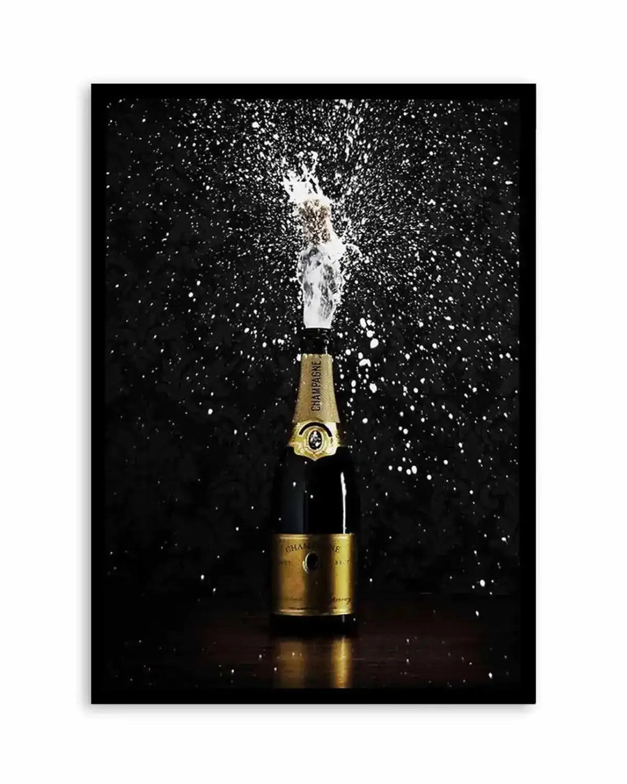 Framed photograph of a champagne bottle popping with a burst of white foam and droplets against a dark, textured background, with a black frame and white border. The champagne bottle is dark green with a gold label, and the cork is mid-air, surrounded by the spray.