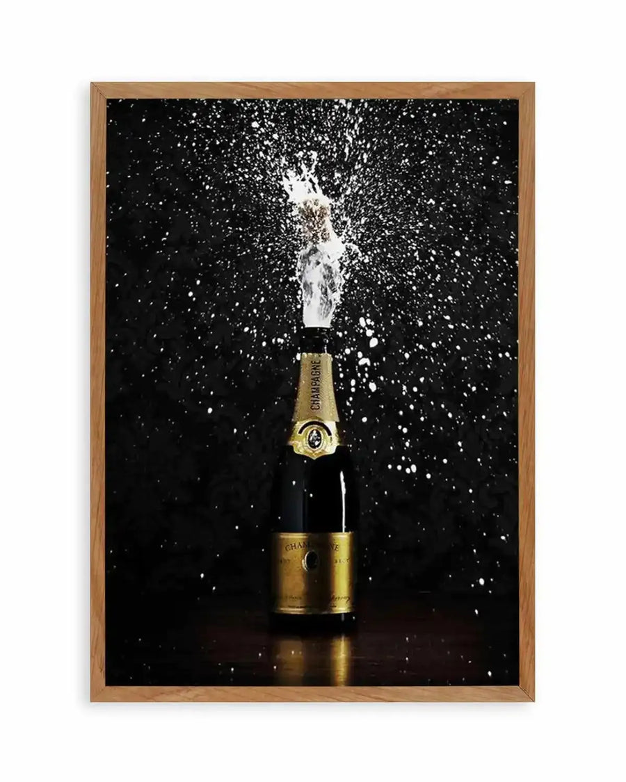 Framed photograph of a champagne bottle popping, with a burst of white foam and sparkling droplets against a dark black background, displayed in a natural oak frame with white border. The bottle is dark green with a gold label, and the cork is mid-air, surrounded by the effervescent spray.