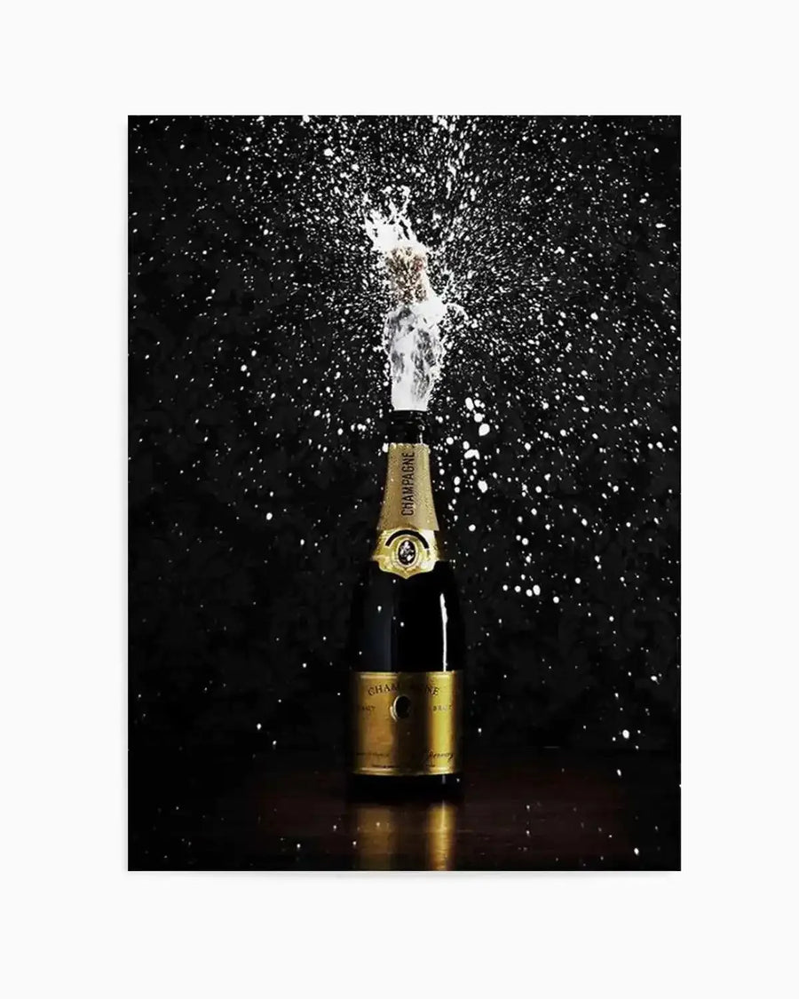 Framed black and white photograph of a champagne bottle popping, with the cork flying upwards and champagne spraying outwards in a dramatic burst against a dark background. The bottle is gold and black, with the word "CHAMPAGNE" visible on the label. The composition captures the dynamic motion of the spray, creating a celebratory and energetic mood.