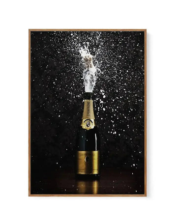 Framed canvas art print featuring a champagne bottle with gold foil and black label against a black background with white champagne spray erupting from the open top. The composition captures dynamic splashing liquid frozen in mid-air, creating a celebratory effect. Natural wood-toned frame with white border surrounding the photograph-style artwork.
