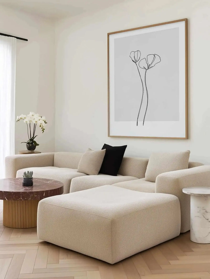 Minimalist line drawing art print displayed in a natural wood frame on a cream wall above a curved beige upholstered sofa. The artwork features two delicate poppy flowers with thin, elongated stems rendered in simple black ink lines on a light gray background, with a white border. The print is shown in a styled living room setting with a burgundy marble side table, white marble cylinder accent table, cream textured cushions including one black pillow, and a potted white orchid by the window.
