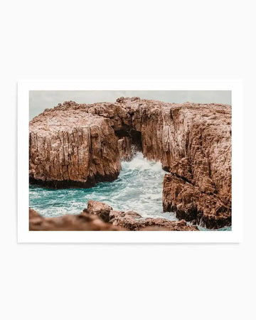 Photography art print featuring a dramatic coastal scene with a natural rock archway over turquoise ocean water. The composition captures waves crashing against the rugged, reddish-brown cliffs and through the arch, creating white foam. The sky is overcast, and the overall mood is powerful and serene.