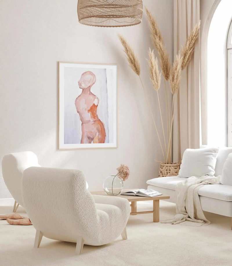 Lifestyle shot of a minimalist living room featuring a framed art print above two cream boucle armchairs. The print depicts an abstract female torso in muted peach and terracotta tones against a soft white background, with a natural wood frame and white border. The room includes a white sofa with a cream throw, a wooden coffee table with a glass vase and dried flowers, and a large vase of pampas grass.