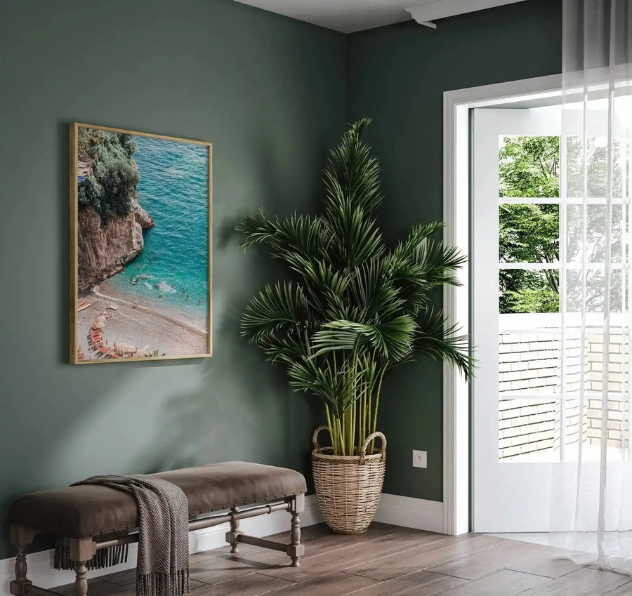 Lifestyle shot of a framed art print displayed on a dark green wall in a corner of a room. The print features a vibrant aerial photograph of a secluded beach with turquoise water, a rocky cliff covered in lush green foliage, and a sandy shore lined with red lounge chairs and white umbrellas. The room includes a brown leather bench with a textured grey throw, a large potted palm plant in a woven basket, and a white door leading to a balcony with a view of trees and a brick wall.