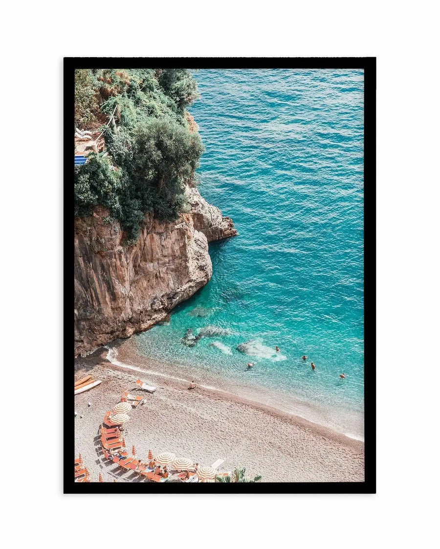 Framed art print featuring an overhead aerial photograph of a Mediterranean beach cove. The composition shows a small sandy beach with striped umbrellas and loungers in coral and cream tones, crystal-clear turquoise water with swimmers, and a dramatic rocky cliff face on the left covered in green vegetation. The photograph captures the gradient from pale sandy beach to shallow aquamarine water to deeper blue sea. Black frame with white border.