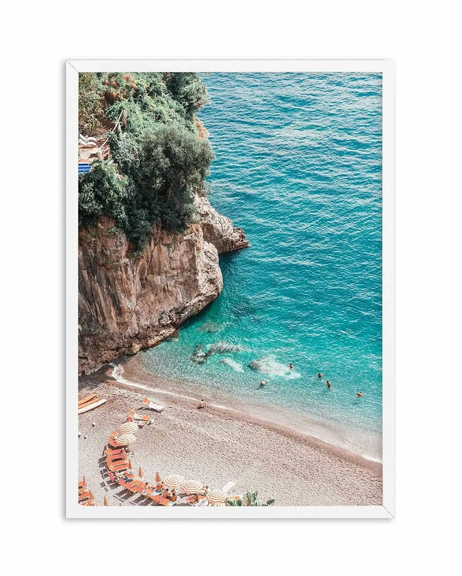 Framed photography art print of a vibrant coastal beach scene with a white frame and white border. The overhead composition features a sandy beach curving around the base of a rocky, tree-covered cliff, with clear turquoise water where several people are swimming. Orange and white striped beach umbrellas and sun loungers are arranged on the sand.