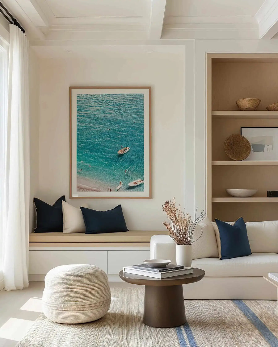 Lifestyle shot of a minimalist living room featuring a large framed art print above a built-in bench. The print, titled "Positano Sands," is a vibrant aerial photograph of a beach with turquoise water, a sandy shore, and small boats, framed in natural wood with a white border. The room includes a white sofa with navy and cream pillows, a round coffee table, and a woven ottoman.