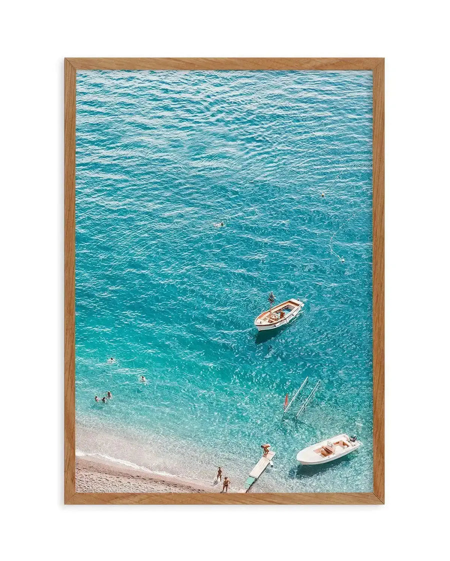 Framed aerial photograph of a vibrant blue ocean with a sandy beach and people swimming and sunbathing, displayed in a natural oak frame with white border. The composition captures the clear turquoise water, revealing the sandy bottom near the shore where several people are wading and swimming. Two small boats are anchored in the water, and a wooden pier extends from the beach with people on it.