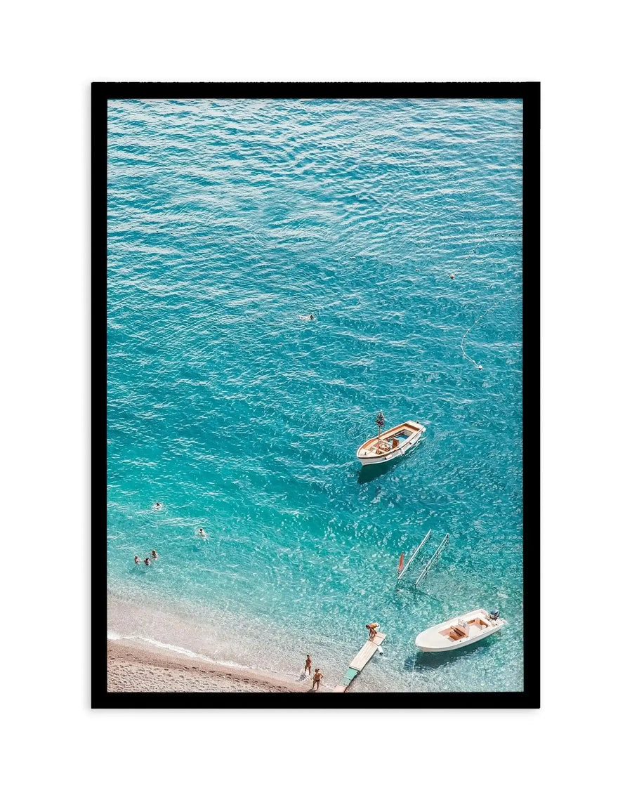 Aerial photograph framed in black showing a crystal turquoise Mediterranean beach cove. White sandy shore in foreground with several swimmers wading in shallow water. Three white and tan fishing boats with visible moorings scattered across the transparent aquamarine water. Deeper teal-blue water fills the upper portion of the composition. Art print presented in black frame with white border.