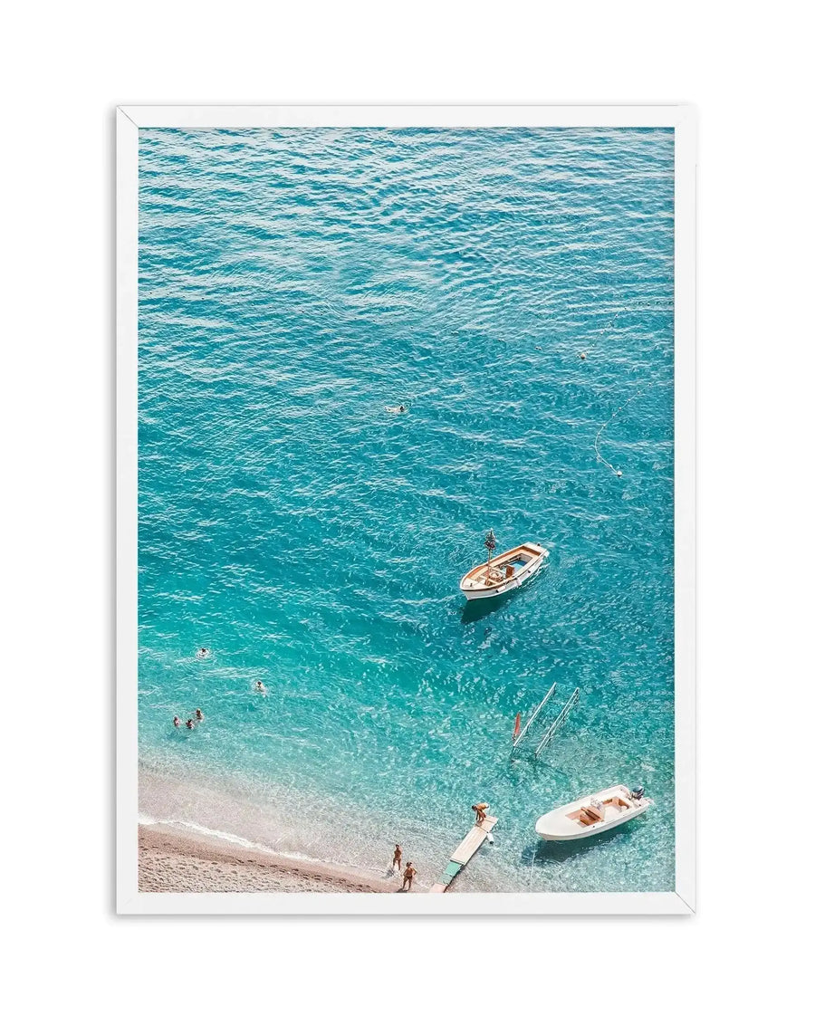 Aerial view art print showing crystal turquoise Mediterranean water with three white fishing boats and swimmers near a sandy beach. Shot from above captures rippling water texture, shallow transparent areas near shore, and scattered figures. White framed print with white border displaying the coastal scene in vibrant teals, soft sand tones, and cream accents. Vertical composition emphasizing the serene seaside landscape.