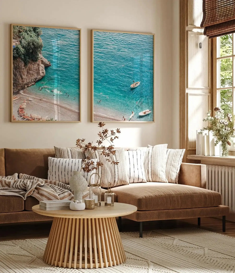 Framed art print displayed on a cream wall above a brown sectional sofa. The print shows an overhead view of a Mediterranean beach cove with turquoise water, sandy shore, scattered sunbathers, and two moored boats. Rocky cliffs with green vegetation frame the left side. The photograph is presented in a light wood frame with white border. Room styled with striped cream and tan pillows, wooden coffee table with fluted base, potted greenery, and natural light from tall windows with woven shades.