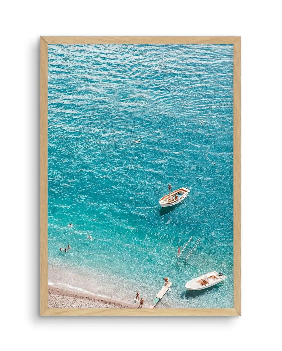 Framed photography art print of a vibrant turquoise ocean scene with a natural wood frame and white border. The aerial view captures a pebbled beach with clear shallow water where people are swimming and standing on a wooden dock. Two small boats are anchored in the deeper blue water.