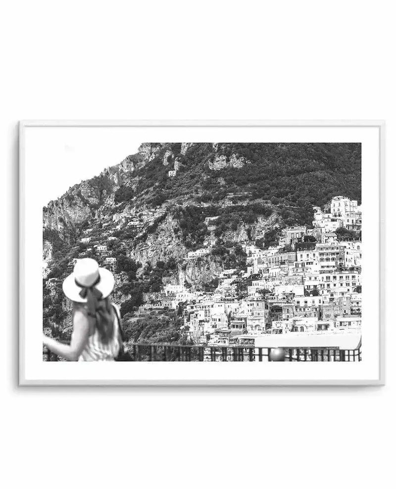 Framed black and white photograph of a coastal town built into a steep mountainside, with a person in a straw hat viewing the scene from a balcony in the foreground. The town's buildings are densely packed, cascading down the mountain towards the bottom right of the frame, with lush vegetation on the upper slopes. The print is displayed in a white frame with a white border.