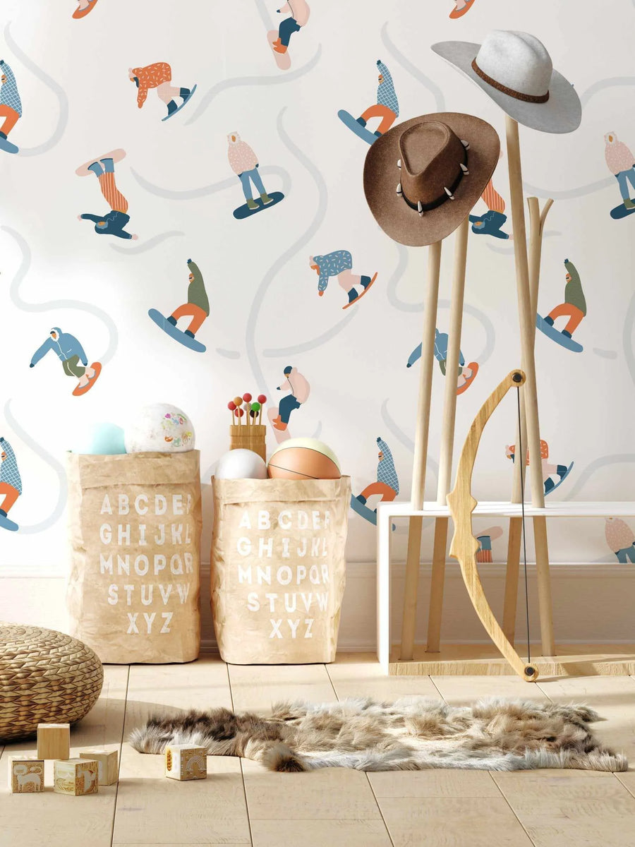 Kids Room:
"Powder Days wallpaper featuring a repeating pattern of playful, illustrated snowboarders in various poses and colourful winter attire on a clean white background with light grey curved lines resembling ski slopes. The wallpaper is displayed in a child's room with light wooden flooring, a woven pouf, wooden alphabet blocks, two brown paper storage bags with white alphabet letters, a faux fur rug, a wooden coat rack with two hats, and a wooden toy bow."