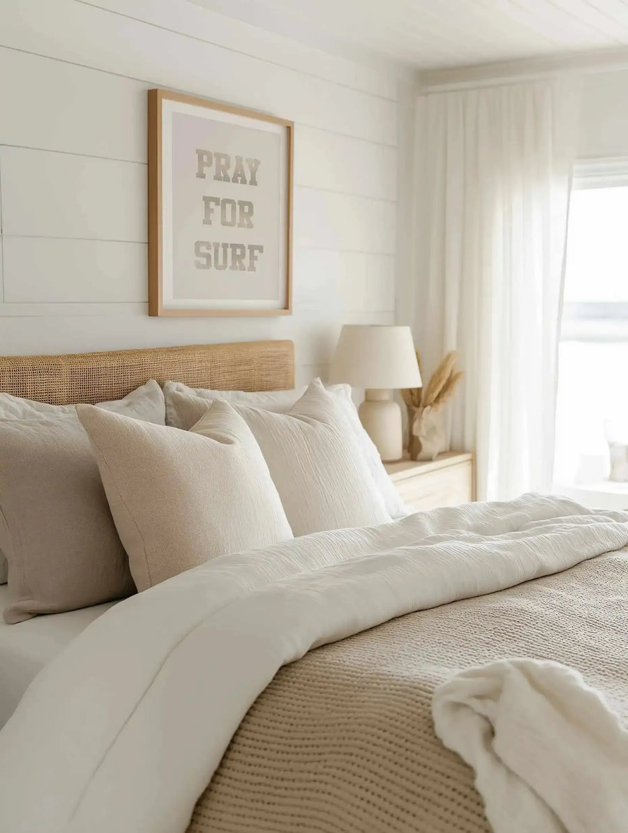 Lifestyle shot of a bedroom featuring a framed art print above a bed with a rattan headboard. The print reads "PRAY FOR SURF" in distressed grey block letters on a light cream background, displayed in a natural wood frame with a white border. The bed is dressed with white and light beige pillows and bedding, and a cream ceramic lamp with a white shade sits on a light wood bedside table next to a vase with dried pampas grass. White sheer curtains hang in the background.
