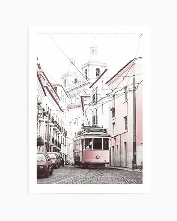 Framed photography art print of a vintage pink tram in a narrow cobblestone street in Lisbon, Portugal. The tram, with "PRAZERES 28" on its destination sign, is turning a corner, surrounded by pastel pink and white buildings with terracotta roofs. In the background, two white church domes rise above the rooftops, and overhead, numerous power lines crisscross the sky. The image has a soft, desaturated, high-key aesthetic with a subtle pink colour cast.