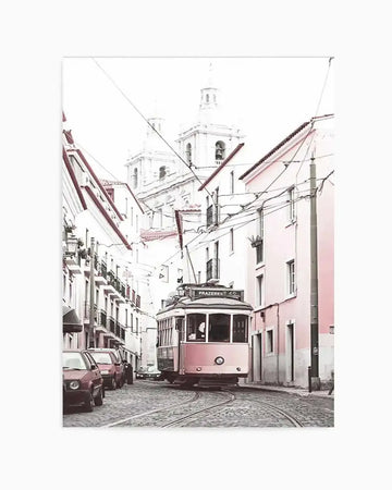 "Photography art print of a street scene in Lisbon, Portugal, featuring a vintage pink tram on cobblestone tracks. The tram is turning a corner, surrounded by buildings with white and pale pink facades, and overhead power lines. In the background, white church steeples rise above the rooftops, under a bright, almost white sky. The image has a soft, slightly desaturated colour palette with a nostalgic mood."