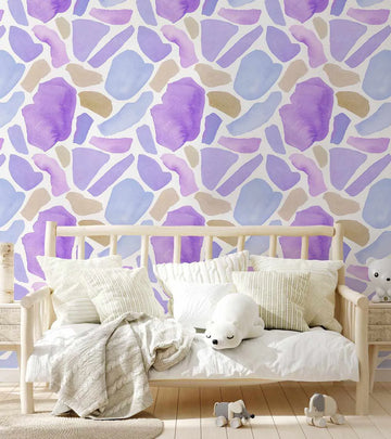 Lifestyle shot of a child's bedroom featuring Provence Terrazzo Wallpaper. The wallpaper has a repeating pattern of irregular, organic shapes resembling terrazzo fragments, rendered in soft watercolour washes of lavender, light purple, pale blue, and warm beige on a white background. The wallpaper is displayed behind a light wood daybed dressed in white bedding and pillows, with a cream cable-knit throw. A white polar bear plush toy and two elephant toys are visible in the foreground.