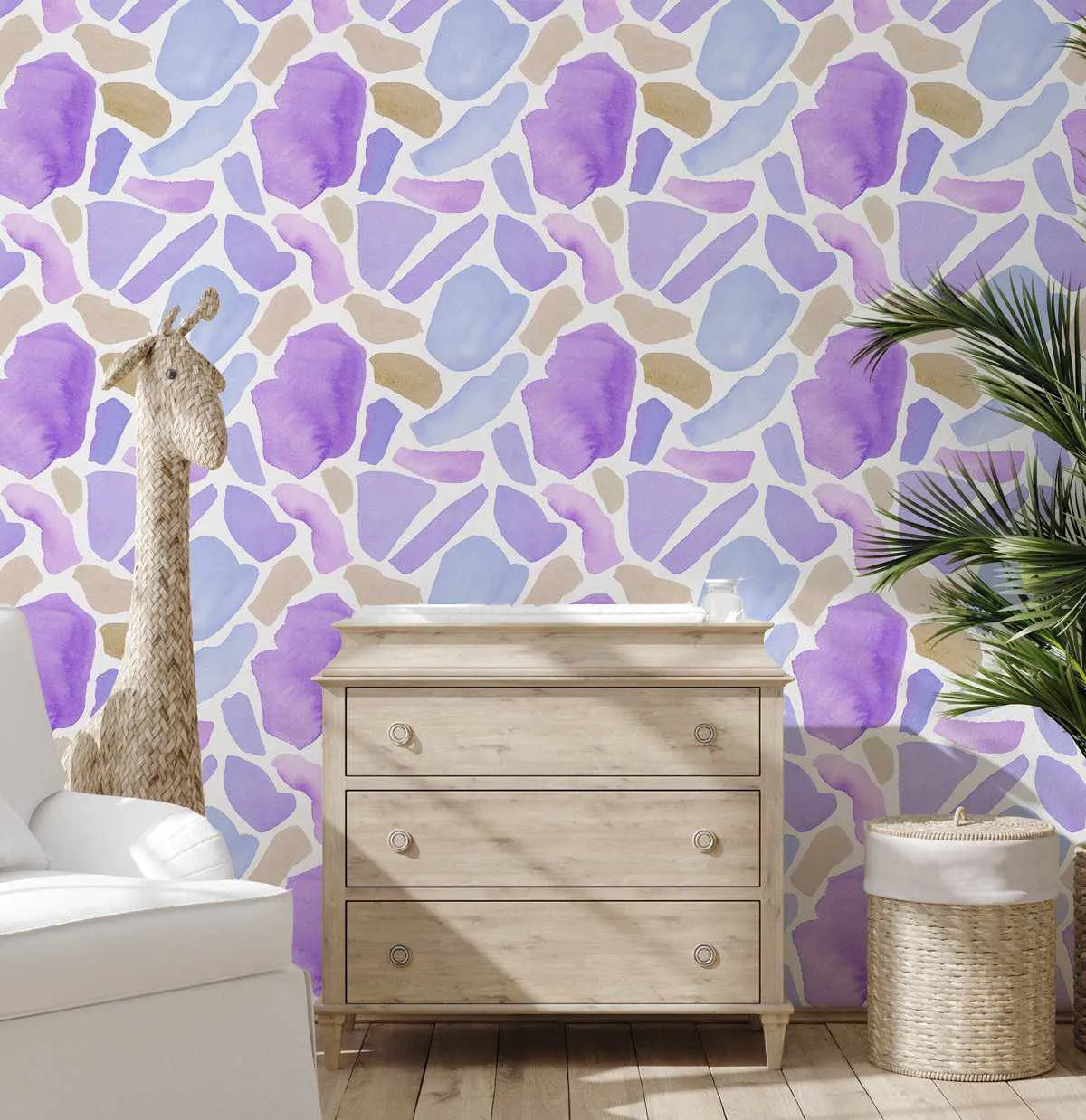 Peel and stick wallpaper featuring a repeating pattern of abstract, organic shapes in varying shades of purple, lavender, light blue, and beige, with a watercolor texture. The wallpaper is displayed in a nursery setting behind a light wood dresser and a white armchair, with a woven giraffe toy and a plant visible.