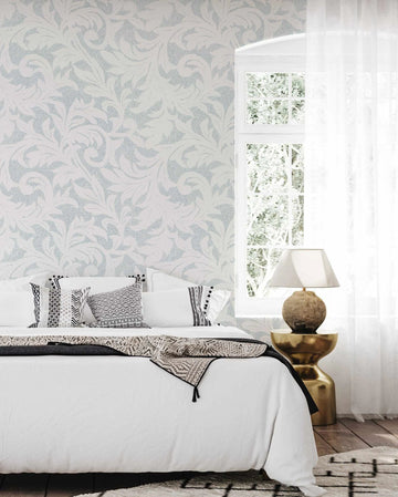 Provence in Ocean wallpaper displayed on a bedroom wall behind a bed with white linens and patterned pillows in black and white. The wallpaper features a repeating design of leafy botanical motifs in soft blue-grey and pale cream tones with a textured, dotted appearance. Tall windows with white frames show greenery outside, and a brass side table with a spherical base and white linen lampshade sits beside the bed. A woven throw blanket drapes across the foot of the bed.