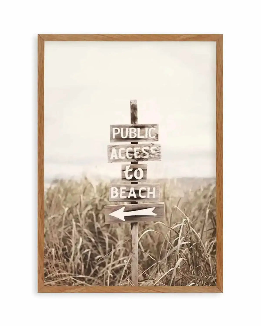 Framed black and white photograph of a rustic wooden signpost on a sandy beach, surrounded by tall, dry grass. The sign has four planks, with "PUBLIC ACCESS to BEACH" painted in white, and a white arrow pointing left on the bottom plank. The photograph has a vintage, sepia-toned quality, displayed in a natural oak frame with white border.