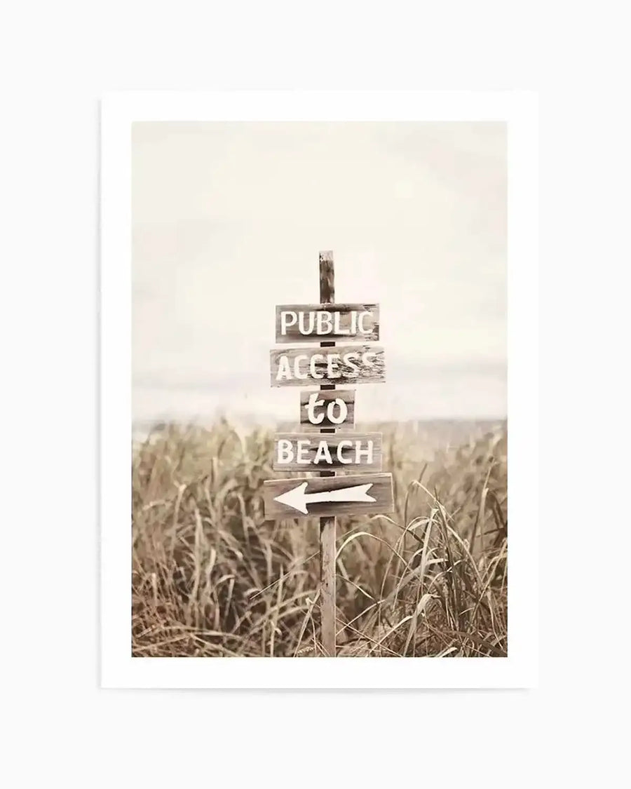 Framed sepia-toned photograph of a weathered wooden signpost on a sandy beach, surrounded by tall, dry dune grasses. The sign has four planks, reading "PUBLIC ACCESS TO BEACH" in white, distressed lettering, with a white arrow pointing left on the bottom plank. The background shows a hazy ocean horizon under an overcast sky.