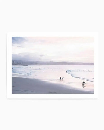 Framed photography art print of a coastal beach scene at dusk with a natural wood frame and white border. The composition features four surfers walking along wet sand at low tide with gentle waves and a soft pink and grey overcast sky stretching to the horizon.