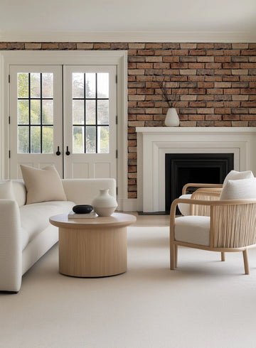 Lifestyle shot of a living room featuring red brick wallpaper on the wall above a white fireplace. The wallpaper has a rustic, textured appearance with varying shades of reddish-brown and dark grey mortar lines. The room includes a cream-colored sofa, a light wood coffee table with decorative vases, and a light wood armchair with a woven back, all set on a light cream carpet.