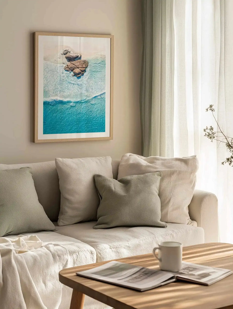 Lifestyle shot of a minimalist living room featuring an aerial photography art print above a cream sofa. The print depicts a coastal scene with two large rock formations on a sandy beach, surrounded by turquoise ocean water and white foamy waves. The print is displayed in a natural oak frame with a white border. The sofa is adorned with cream and sage green cushions and a cream throw, and a wooden coffee table with a magazine and mug is in the foreground.