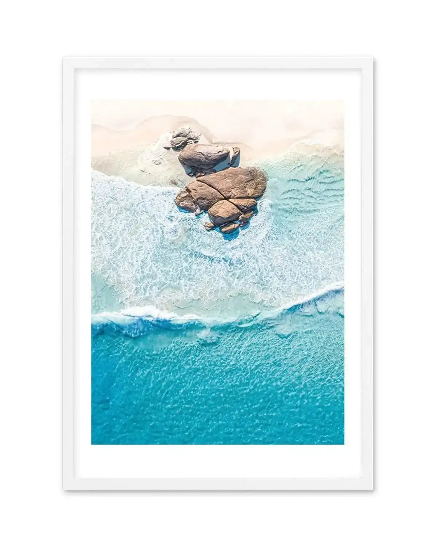 Framed aerial photograph of a coastal beach scene with a white frame and white border. The composition features a white sandy beach with large brown rocks partially submerged in the turquoise ocean, with gentle white waves breaking on the shore.