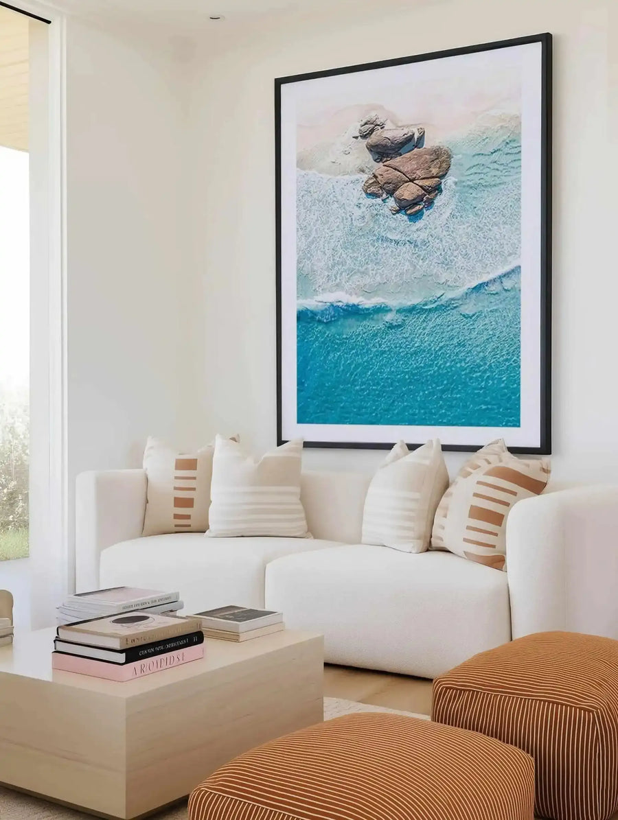 Large framed art print displayed above a curved white sofa in a minimalist living room. The print is an aerial photograph of a beach with large brown rocks on the sand and turquoise water with white waves crashing. The print has a black frame and a white border. The room includes a light wood coffee table with books and two striped orange ottomans.