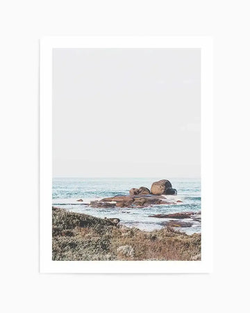 Framed photograph of a coastal scene featuring large, smooth boulders in the ocean with gentle waves and a light, hazy sky. The foreground shows dry, textured coastal vegetation in muted greens and browns. The print is displayed with a white border.
