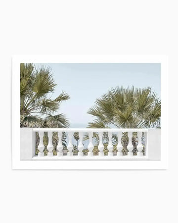 Framed photography art print of a tropical scene with a white balustrade in the foreground. The composition features two large palm trees with green fronds against a pale blue sky, with a glimpse of the ocean in the distance.