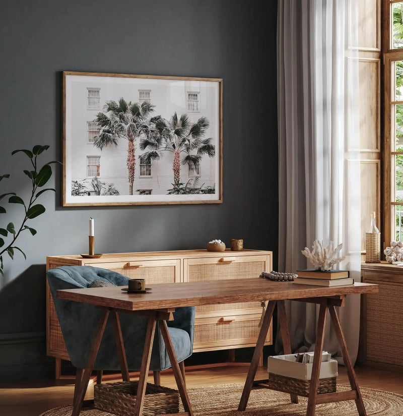 Home Office:
Large framed photography art print displayed above a wooden console table in a home office. The print features a white building with several windows and three palm trees in the foreground, with a natural wood frame and white border. The room includes a wooden desk with a blue upholstered chair, a sheer white curtain, and a window with a view of green foliage.