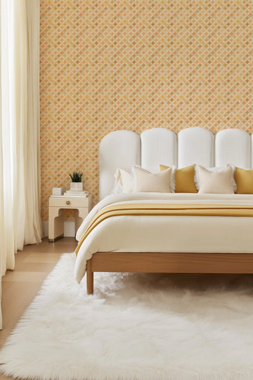 Retro Diamonds wallpaper featuring a repeating pattern of interlocking diamond shapes in a palette of warm beige, muted peach, soft gold, and light brown, installed on a bedroom wall behind a bed. The wallpaper has a subtle textured appearance. The room includes a white upholstered headboard, cream bedding with a mustard yellow throw, a white faux fur rug, and a light wood bedside table with a small plant.